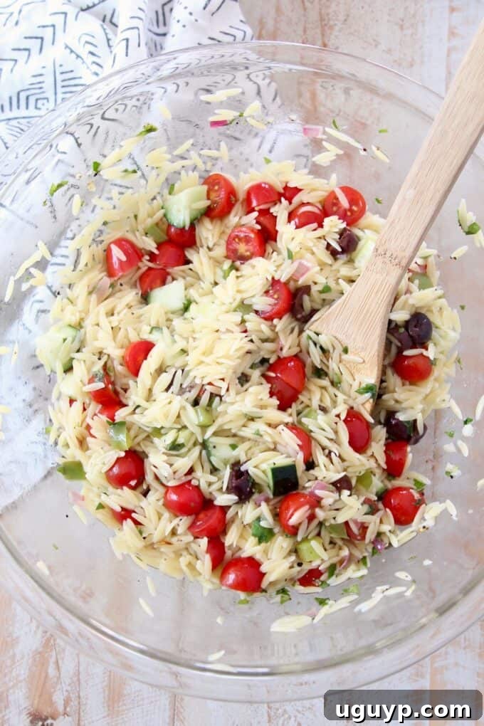 All the Greek Orzo Salad ingredients tossed together in a large glass bowl with a wooden spoon.