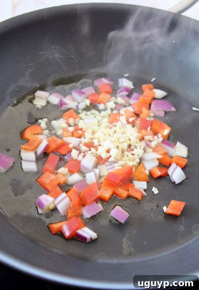 Diced red onion, bell pepper, and garlic sautéing in a hot skillet with olive oil, creating an aromatic base.