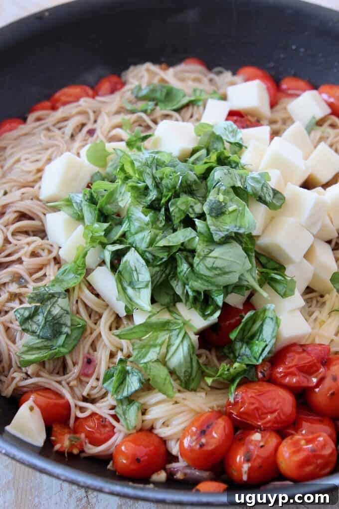 Finished Caprese pasta in the skillet, generously topped with fresh basil leaves and cubes of mozzarella cheese, creating a visually appealing and fragrant dish.