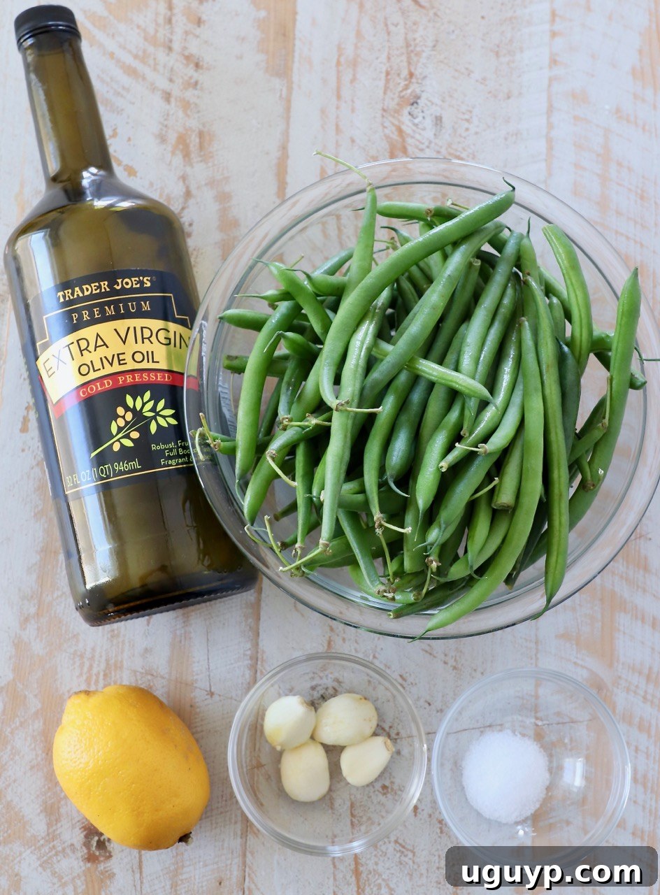 Fresh ingredients laid out on a wooden board: green beans, garlic cloves, a whole lemon, olive oil, and a pinch of salt.
