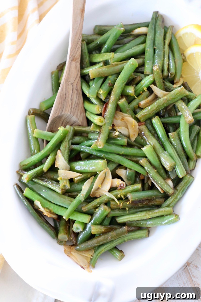 A large white serving dish brimming with vibrant garlic green beans, with a wooden serving spoon nestled within the dish.