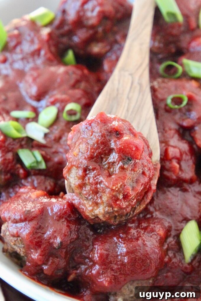 Juicy baked beef meatballs drenched in red wine BBQ sauce in a bowl, with one meatball on a spoon