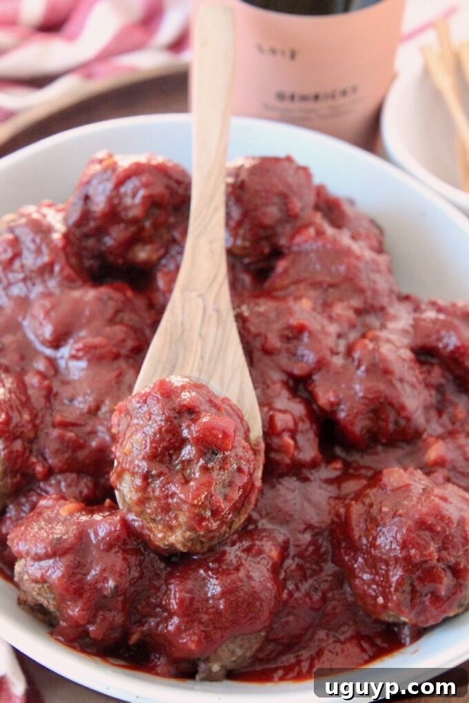 A single baked beef meatball perfectly coated in rich red wine BBQ sauce, held on a spoon over a bowl full of more delicious meatballs.