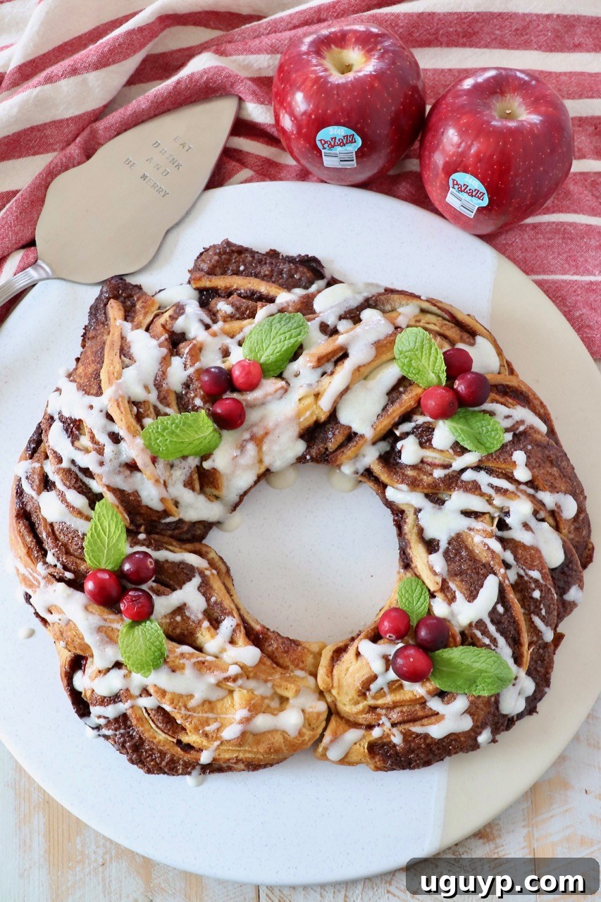 Beautifully baked apple cinnamon roll wreath on a round white serving tray, garnished with fresh cranberries and mint leaves.