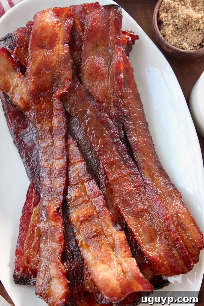Crispy, glossy slices of candied bacon stacked on a white plate, showcasing its irresistible sweet and salty glaze.