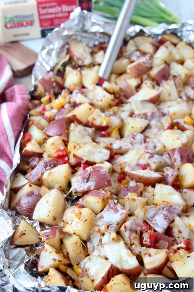 Cubed red potatoes, onions, and peppers grilled in foil, showing melted cheese and a spatula lifting a serving.