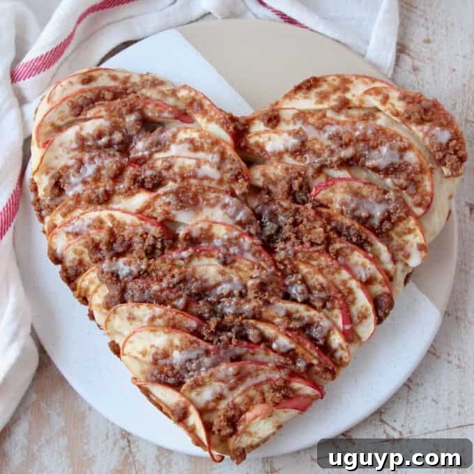 An overhead view of a heart-shaped apple dessert pizza, topped with sliced apples, streusel, and a drizzle of icing.