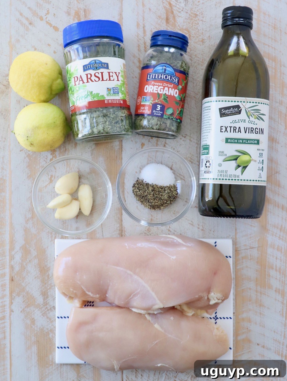 A collection of fresh ingredients laid out on a surface, including olive oil, garlic cloves, lemons, dried oregano, dried parsley, salt, and pepper, ready for making the Greek chicken marinade.