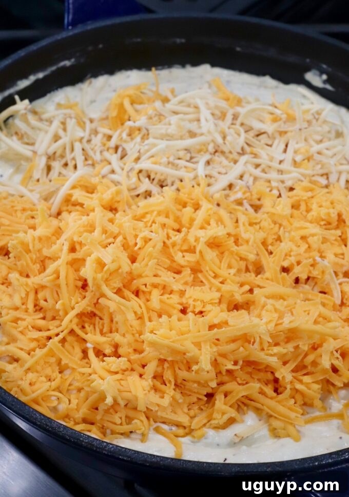 Shredded cheddar and smoked gouda cheese being added to the creamy sauce in a skillet.