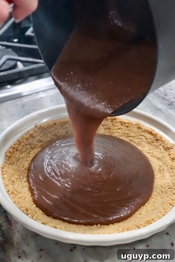 Thick, glossy chocolate filling being poured from a saucepan into the baked graham cracker pie crust.