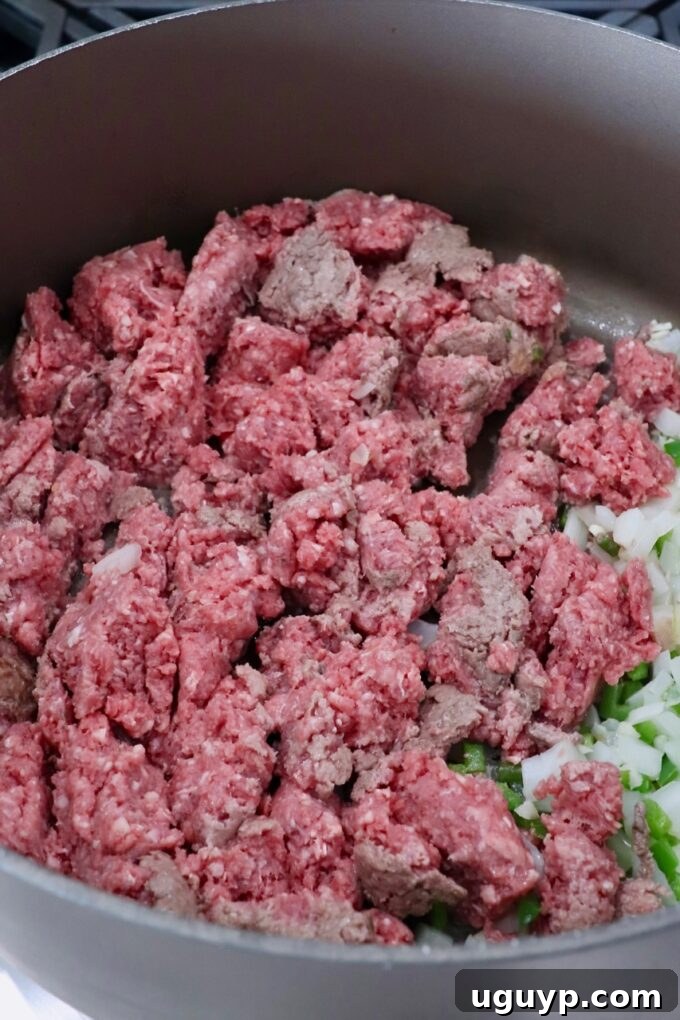 Raw ground beef browning in a large skillet on the stove, alongside sautéed onions, garlic, and jalapenos.