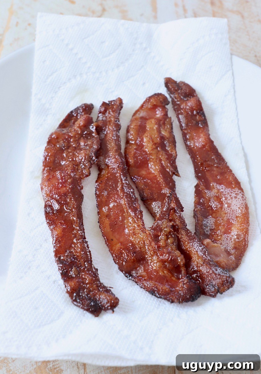 Crispy, golden-brown bacon strips resting on a paper towel-lined plate, absorbing excess grease after being perfectly cooked in the oven.