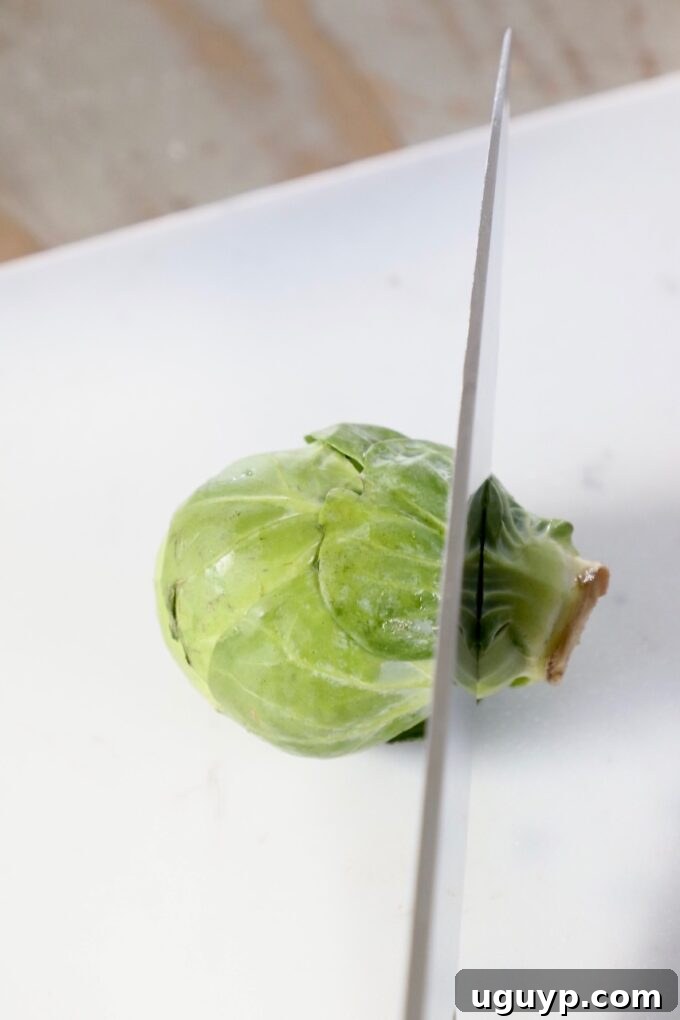 A chef's knife begins to slice into a fresh Brussels sprout on a sturdy wooden cutting board, initiating the shredding process.