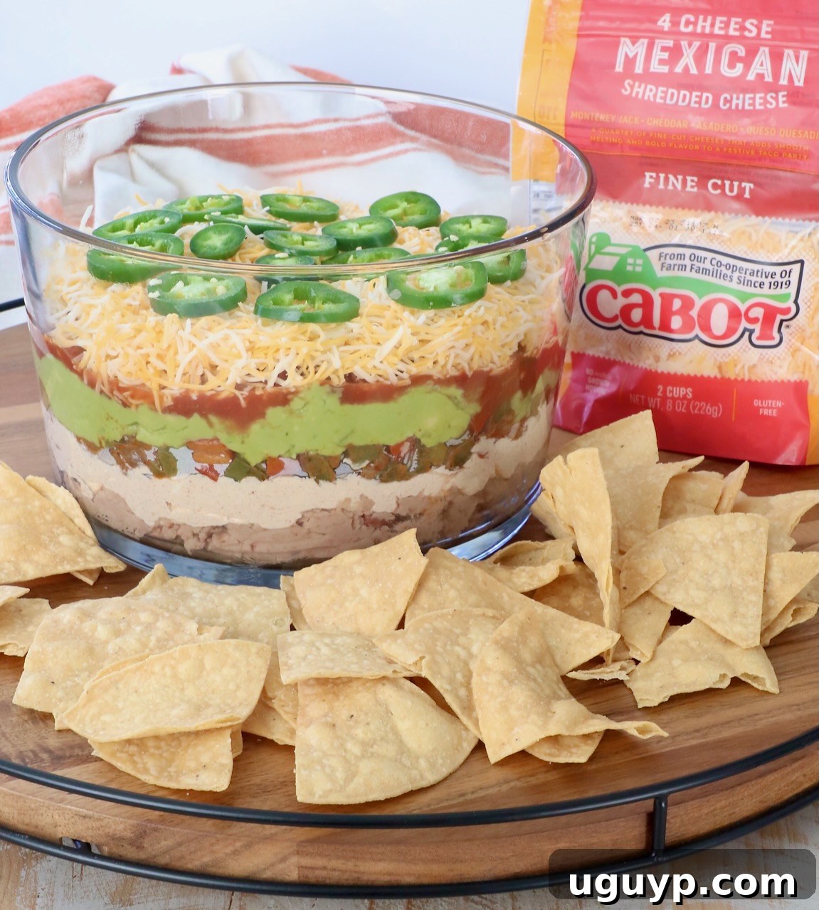 Beautifully presented 7 layer dip in a glass trifle bowl, surrounded by crispy tortilla chips on a rustic serving tray