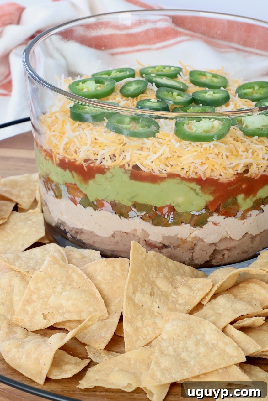 Another view of the 7 layer dip in a glass trifle bowl, served on a tray with tortilla chips, inviting guests to dig in