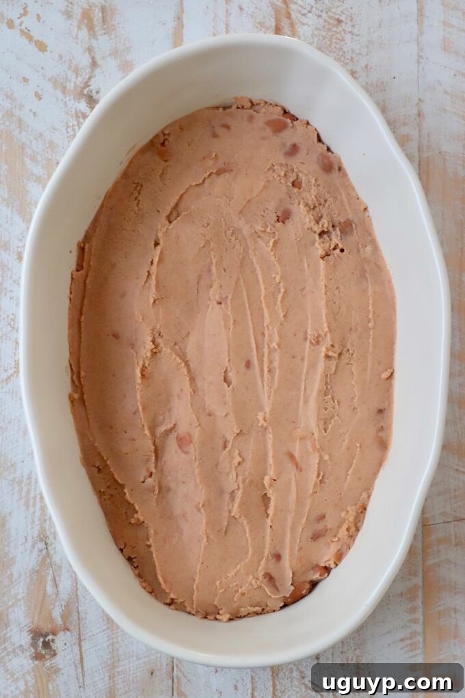 Refried beans spread evenly in a casserole dish, forming the first layer