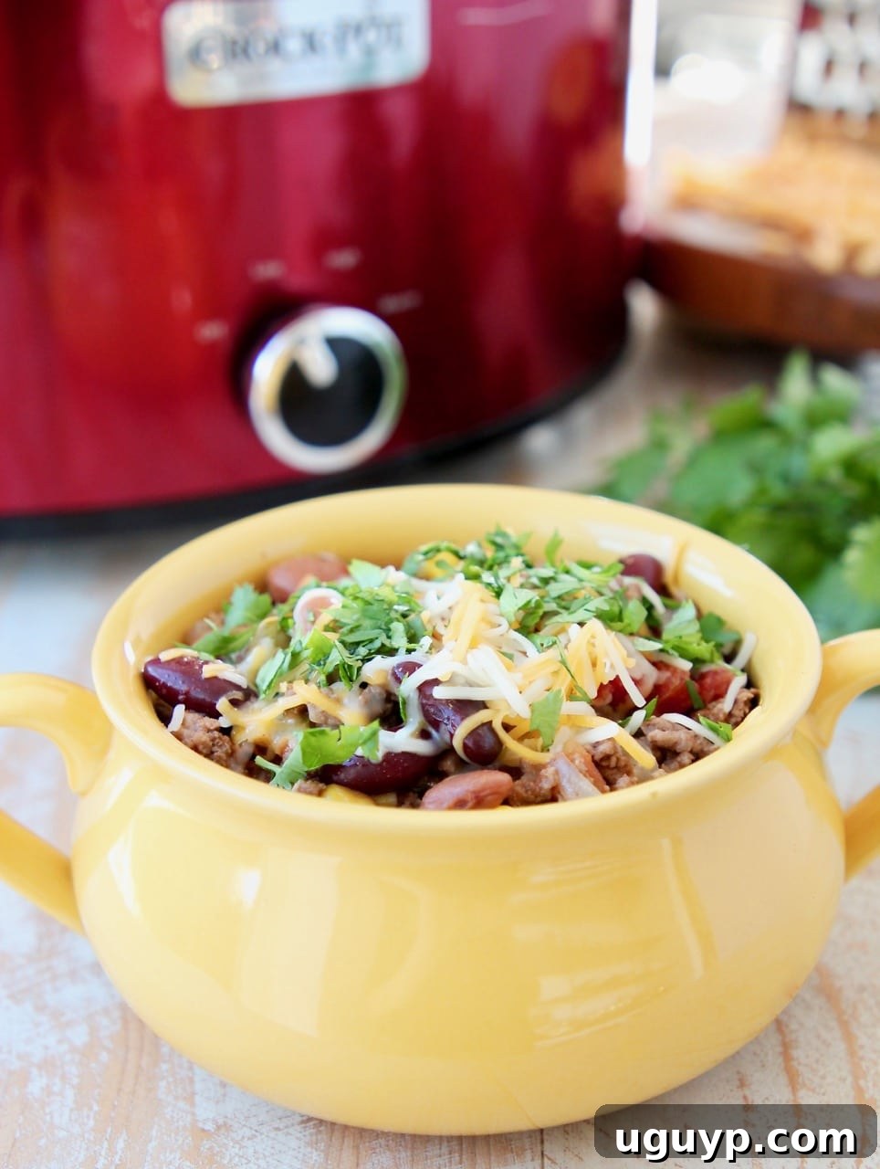 A close-up of a yellow crock filled with Slow Cooker Taco Soup, topped with cheddar cheese and fresh cilantro, alongside a red slow cooker in the background.