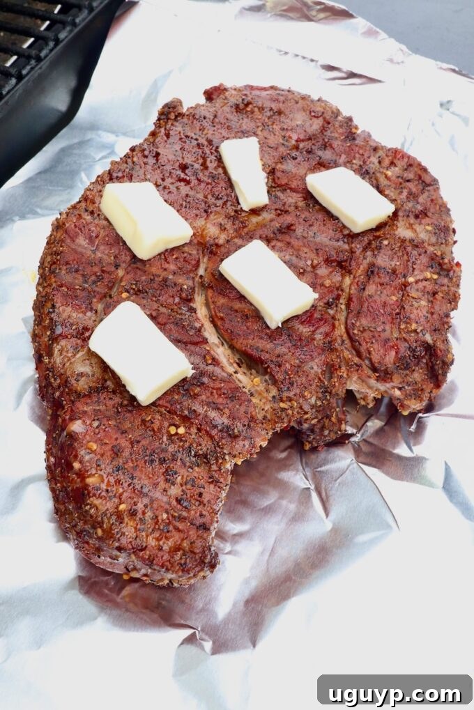 Smoked chuck roast, already seasoned and partially cooked, topped with pats of butter on a piece of foil, ready for wrapping.