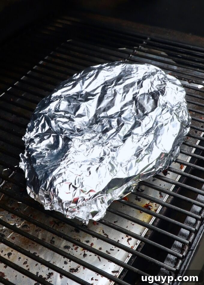 Chuck roast wrapped tightly in foil on the smoker, undergoing the 'Texas Crutch' to enhance tenderness and moisture.