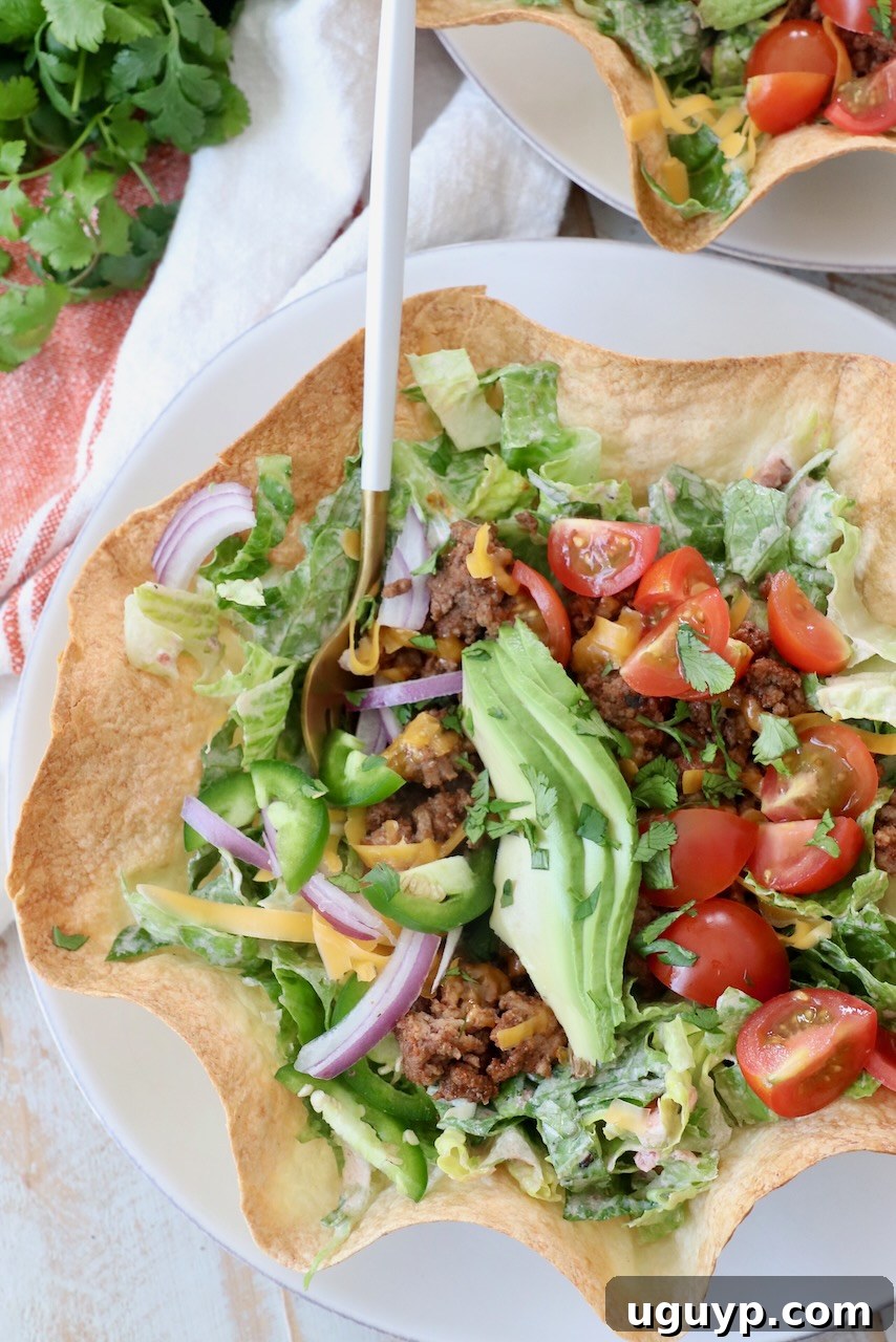 A close-up shot of a fork digging into a taco salad served in a crispy tortilla shell, showcasing the layers of meat, cheese, lettuce, and avocado.