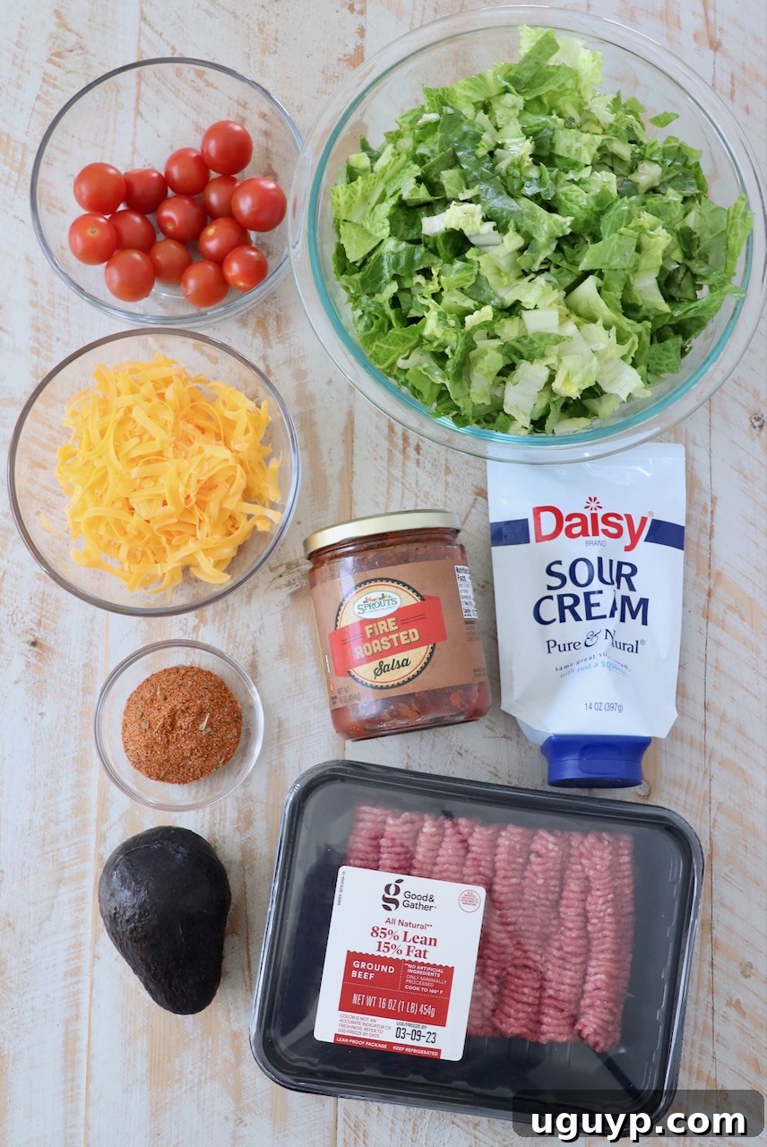A selection of fresh ingredients laid out on a white wooden board, including shredded lettuce, cherry tomatoes, avocado, ground beef, cheese, and a bowl of salsa and sour cream, ready for making taco salad.