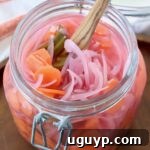 A close-up of vibrant Mexican pickled onions, carrots, and jalapenos, neatly arranged in a large glass mason jar, showcasing their colorful appeal and readiness for enjoyment.