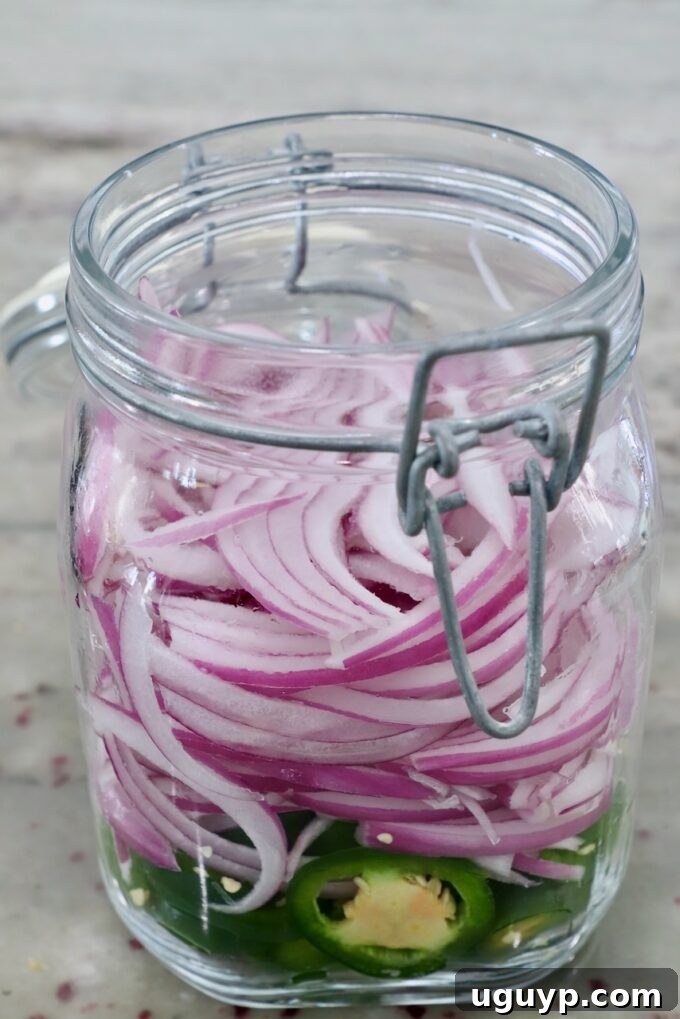 A large glass jar holds a vibrant mix of thinly sliced red onions and jalapenos, awaiting the addition of the pickling liquid to complete the escabeche.