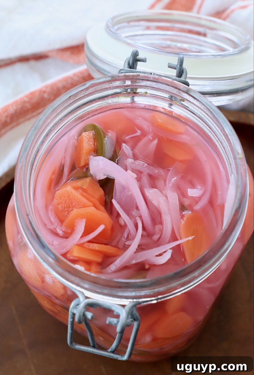 A large glass jar filled with vibrant Mexican Pickled Vegetables, including carrots, onions, and jalapenos, with the lid ajar, inviting viewers to savor its fresh taste.