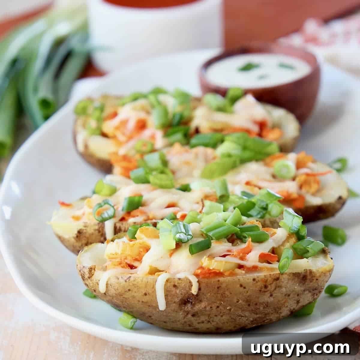 Crispy buffalo chicken potato skins piled on a white plate, garnished with green onions, with bowls of buffalo sauce and ranch in the background.