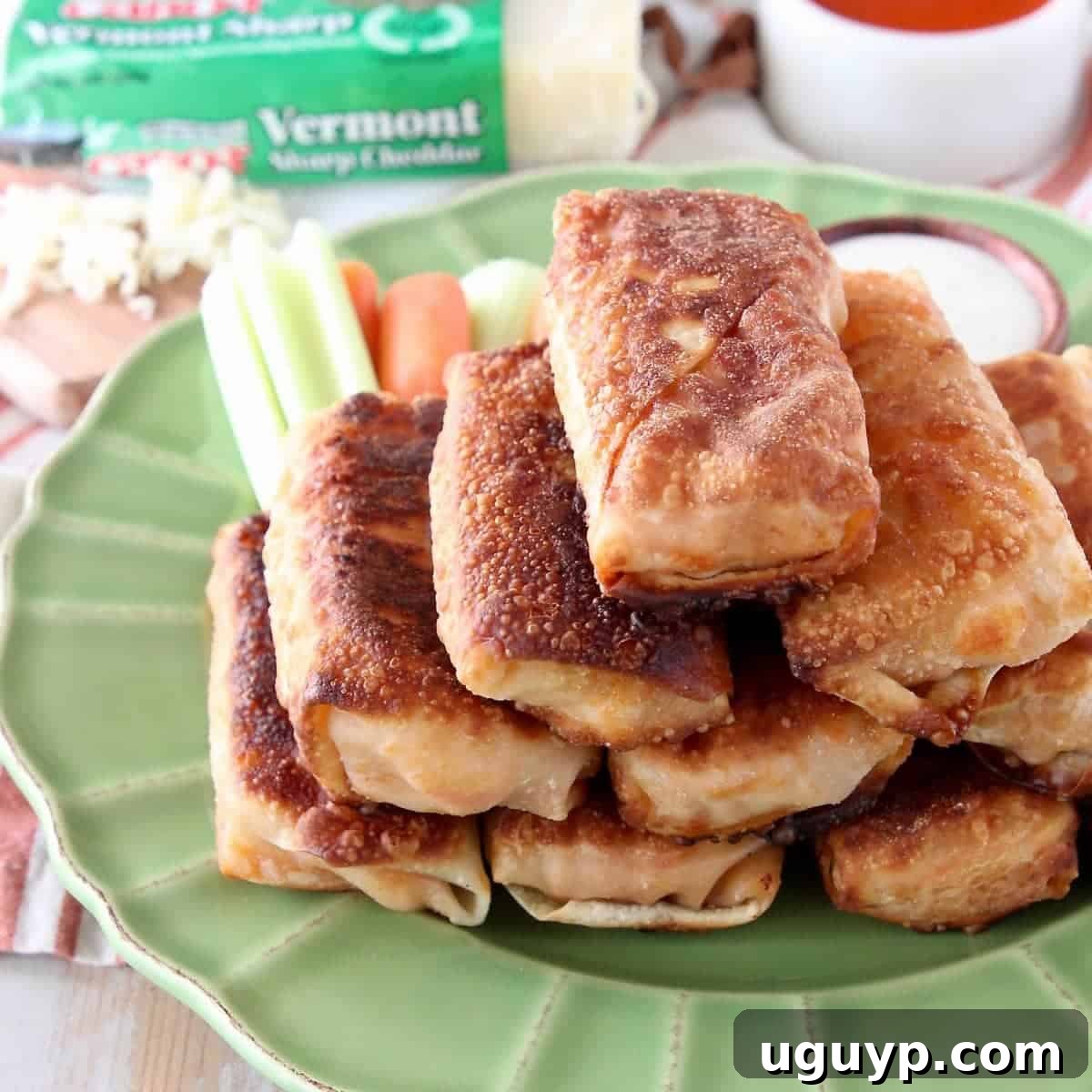 Golden-fried buffalo chicken egg rolls stacked on a green plate, accompanied by crisp celery, carrots, and a small bowl of ranch dressing.