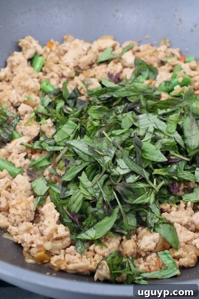 Fresh Thai basil leaves stirred into cooked ground chicken and vegetables in a large pan, infusing the dish with its signature aroma and flavor.