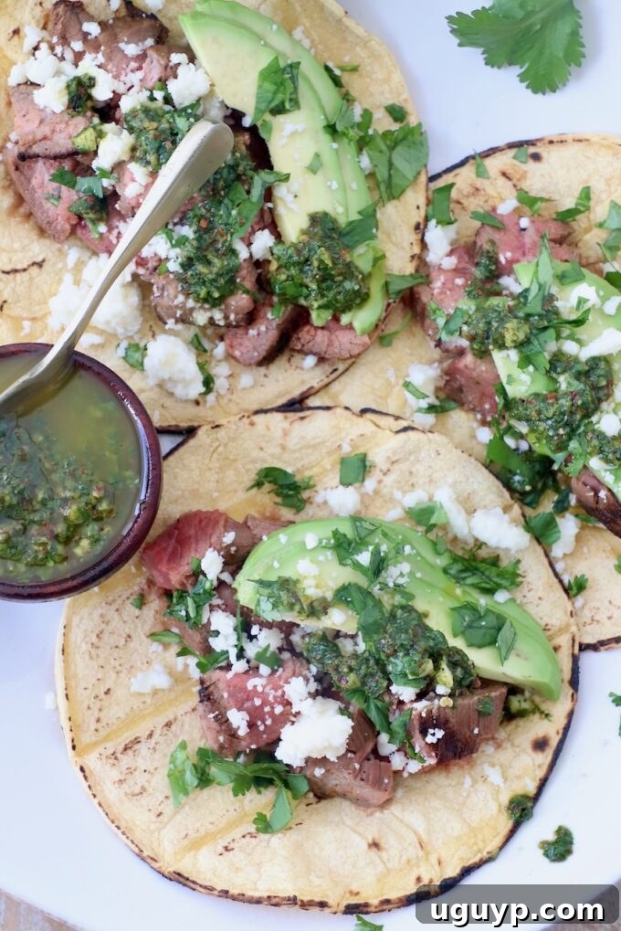 three tacos on white plate with a small bowl of chimichurri sauce on the side