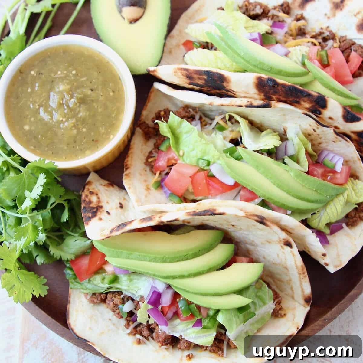Three ground beef tacos on wood tray with avocado and salsa
