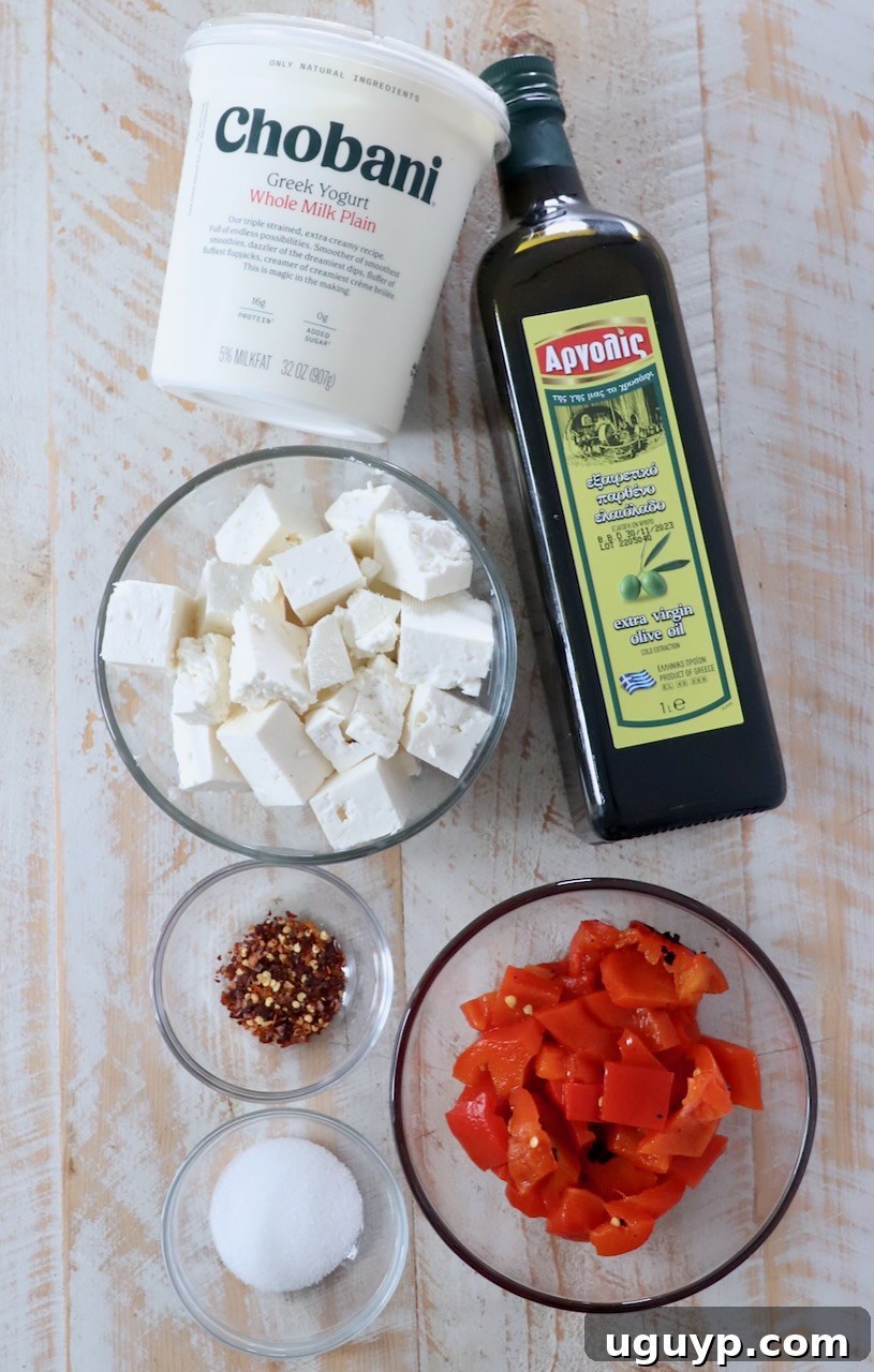 Ingredients laid out for making spicy feta dip, including a block of feta cheese, roasted red peppers, Greek yogurt, olive oil, and red pepper flakes.