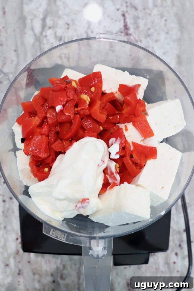 Feta cheese, Greek yogurt, and roasted red peppers are placed inside a food processor, ready for blending into a creamy dip.