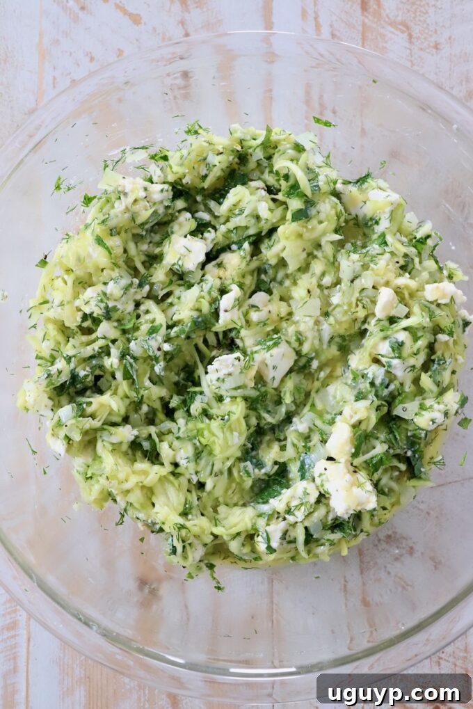 Greek zucchini pie filling mixed together thoroughly in a large glass bowl, showing the vibrant herbs and feta.