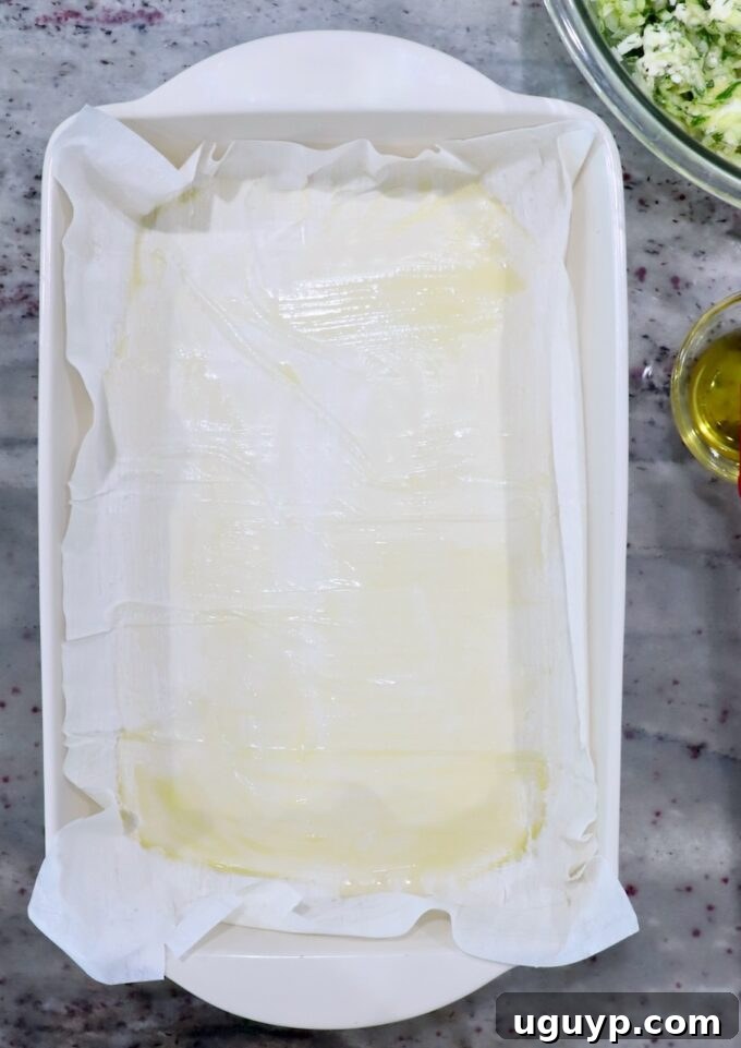 The first sheet of phyllo dough brushed with olive oil in a baking dish, preparing the base for the Greek zucchini pie.