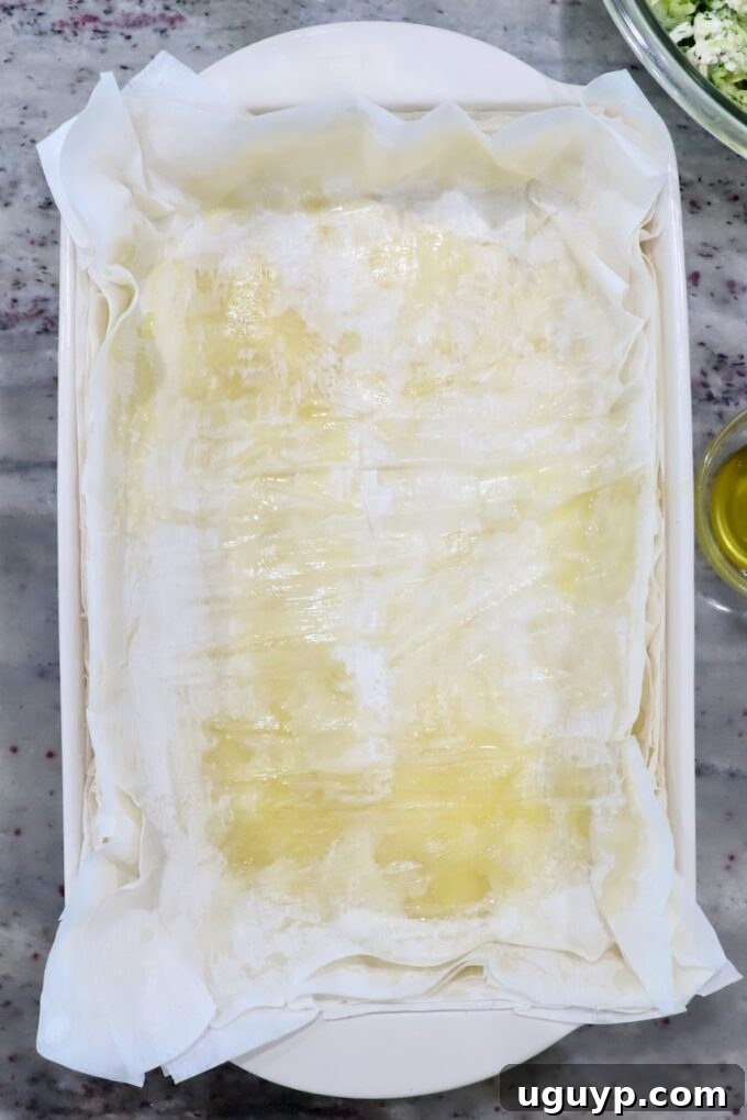 Multiple sheets of phyllo dough layered one on top of the other in a baking dish, each brushed with olive oil, forming the base of the pie.