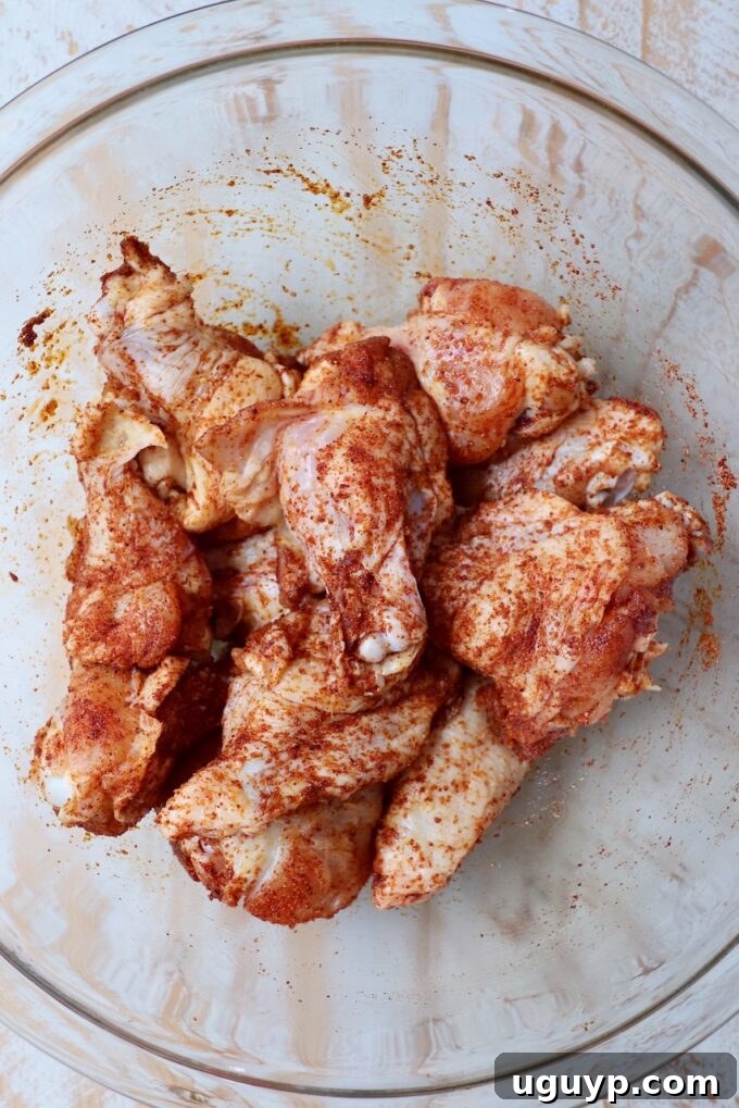 Chicken wings generously coated with dry rub seasoning in a clear glass bowl, ready for the grill.