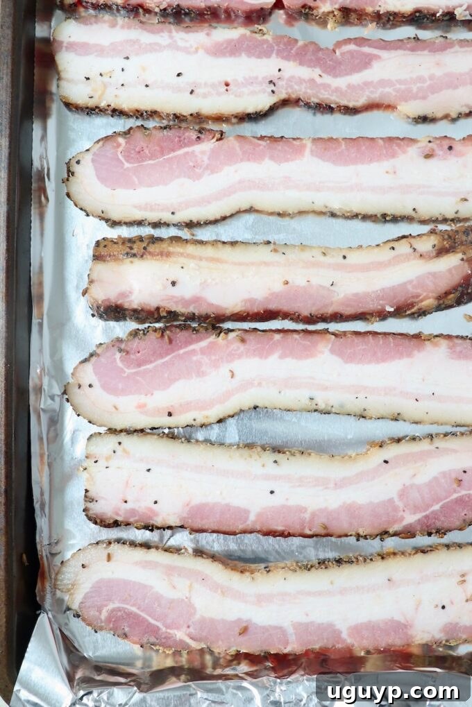 Neatly sliced bacon laid out in a single layer on a foil-lined baking sheet, prepared for baking.