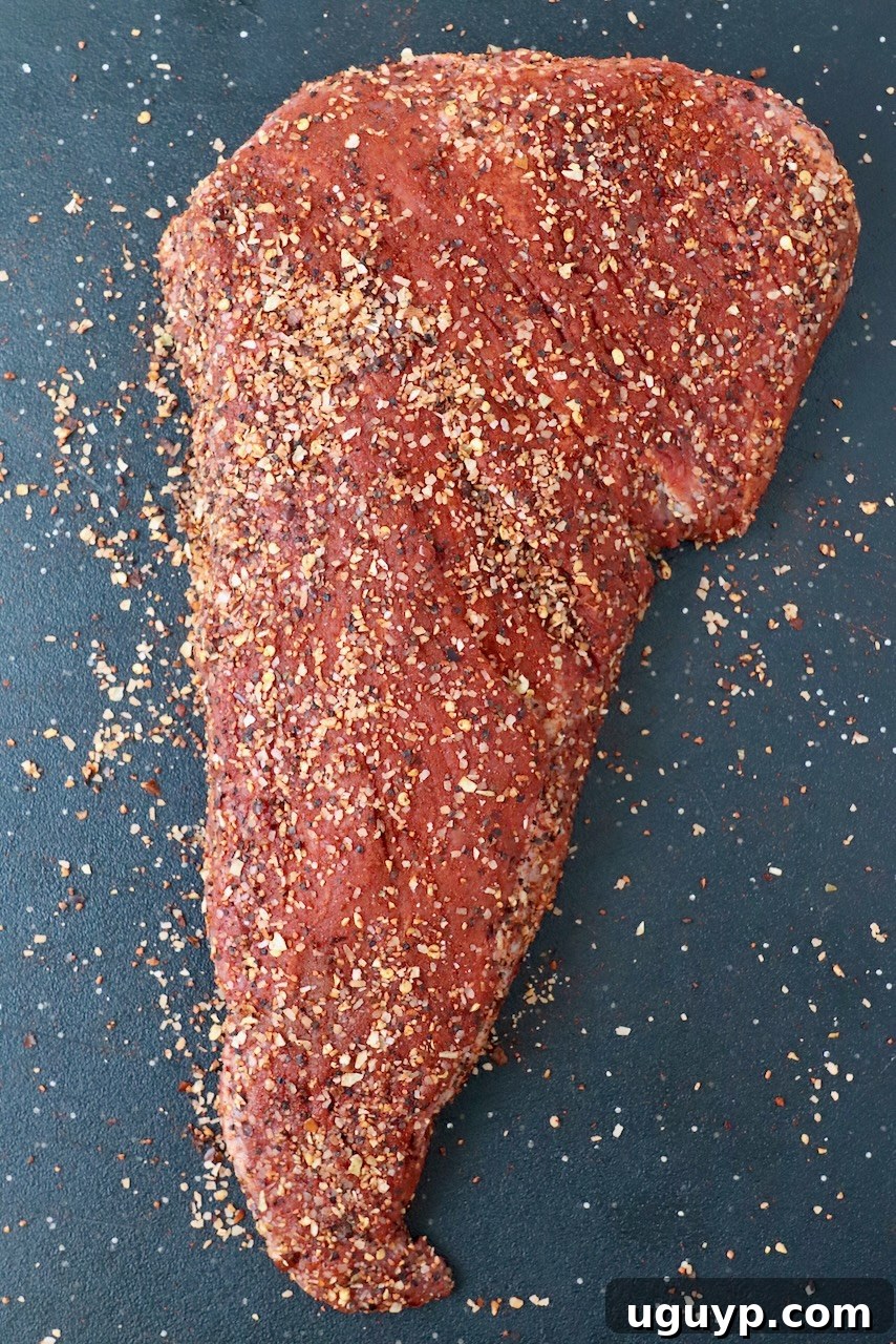 Uncooked tri tip roast generously seasoned with steak rub on a cutting board, ready for the smoker.