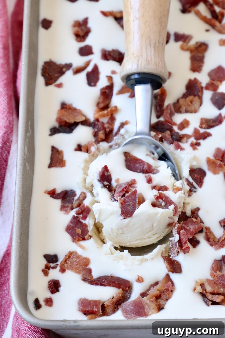Scooped maple bacon ice cream in a metal container, garnished with candied bacon pieces and an ice cream scoop