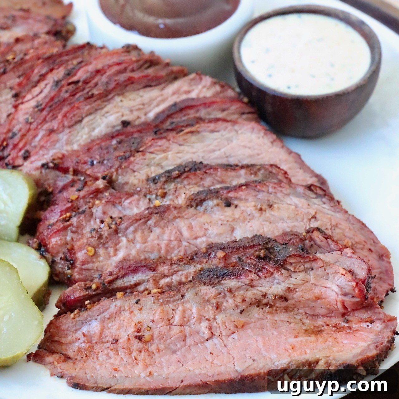 Perfectly sliced smoked tri-tip steak on a plate, accompanied by small bowls of various BBQ sauces, including Alabama White Sauce.