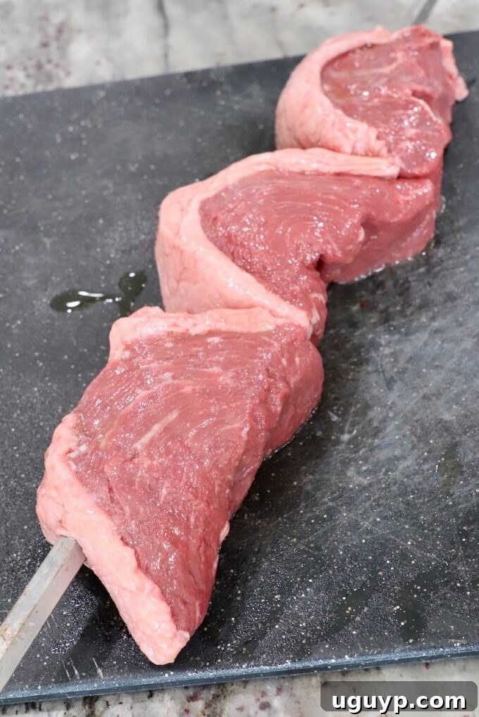 Three C-shaped picanha steaks skewered on a rotisserie rod, resting on a cutting board.