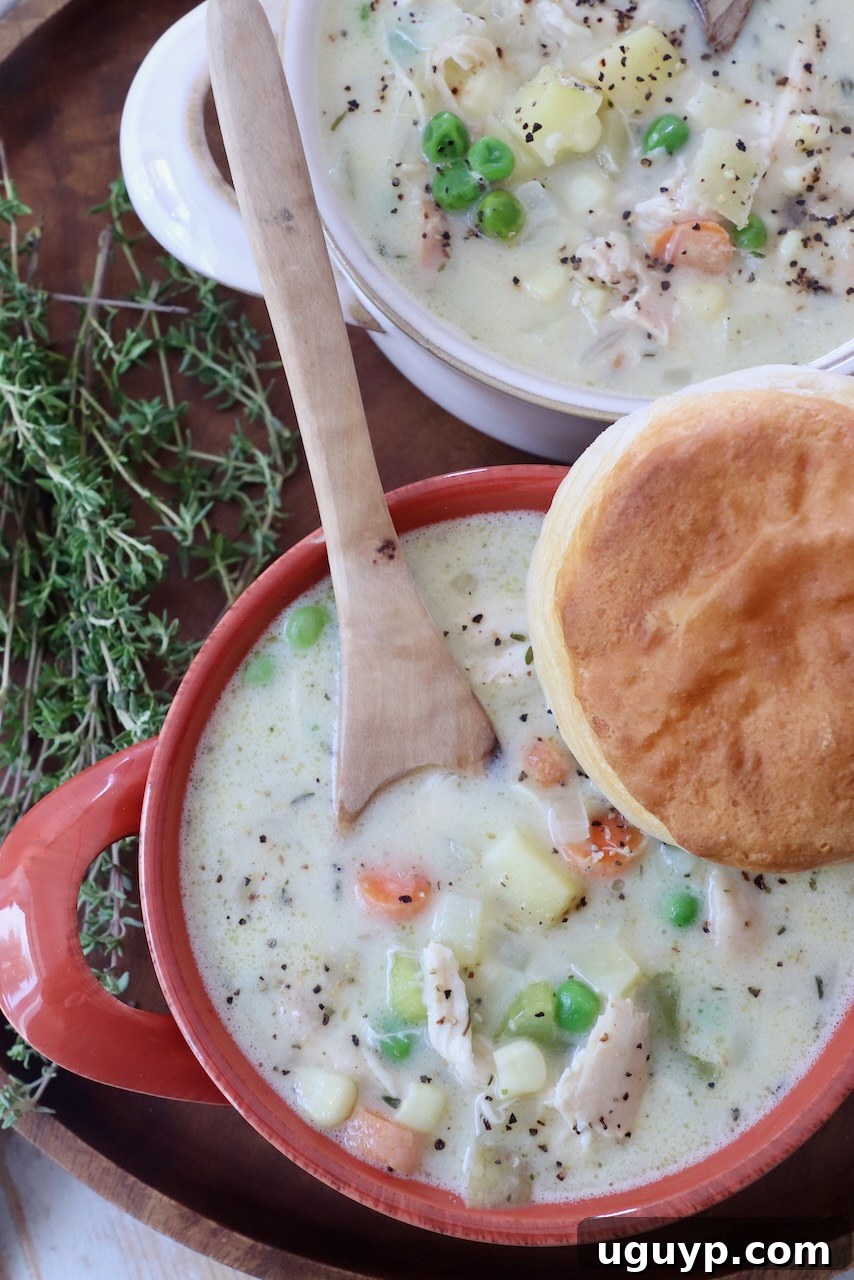 Hearty chicken pot pie soup in a bowl with a wooden spoon and a biscuit for dipping.