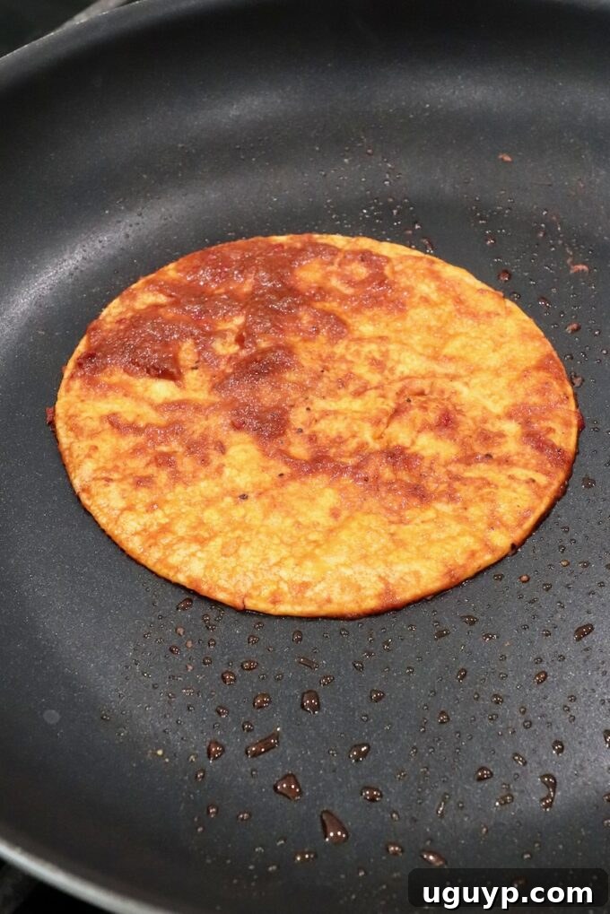 A corn tortilla being dipped into red birria consomé in a hot skillet, ready for assembly.