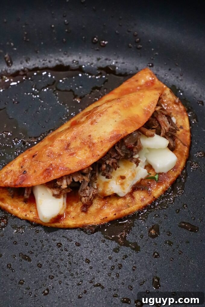 A quesabirria taco, filled with melted cheese and birria beef, cooking in a skillet until golden brown and crispy.