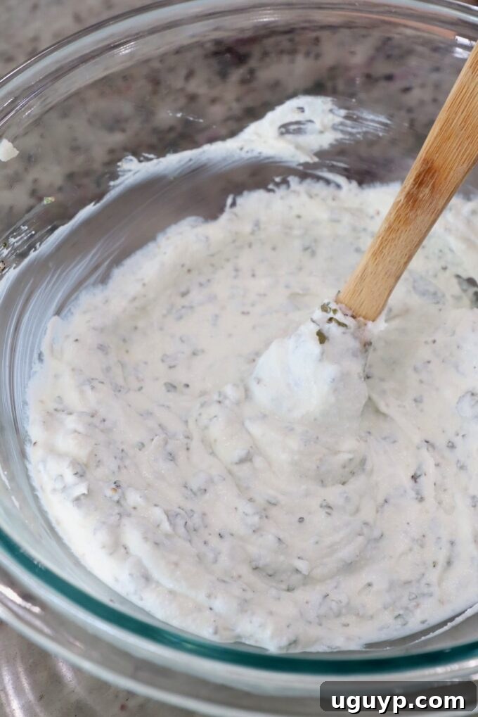 Creamy ricotta cheese, beautifully seasoned with dried basil, parsley, oregano, salt, and pepper, mixed thoroughly in a glass bowl with a spatula, creating a vibrant, herb-infused topping.
