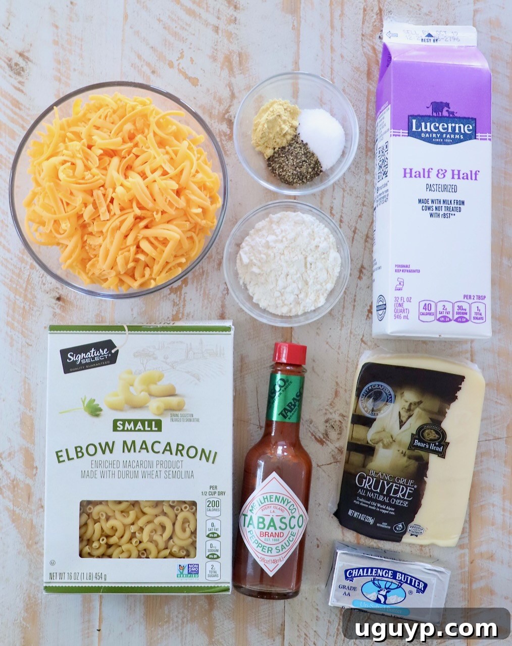 A collection of fresh ingredients for mac and cheese laid out on a rustic white wooden board, including blocks of cheddar and swiss cheese, macaroni noodles, butter, and seasonings.