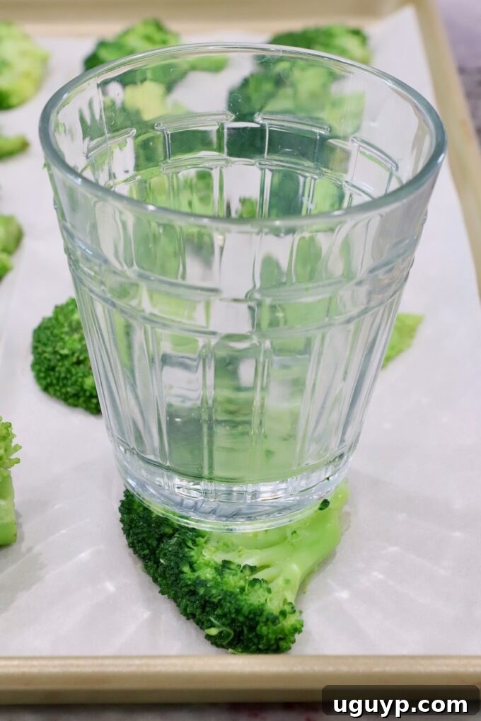 A glass cup pressing down on a piece of steamed broccoli on a baking sheet to smash it flat